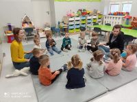 Grupa dzieci oraz dwie opiekunki siedzą w kółku na dywanie i grają w grę